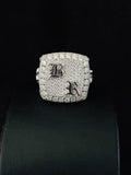 A square-shaped silver ring heavily paved with diamond, a gothic letter 'B' on one side and a gothic letter 'K' on the other