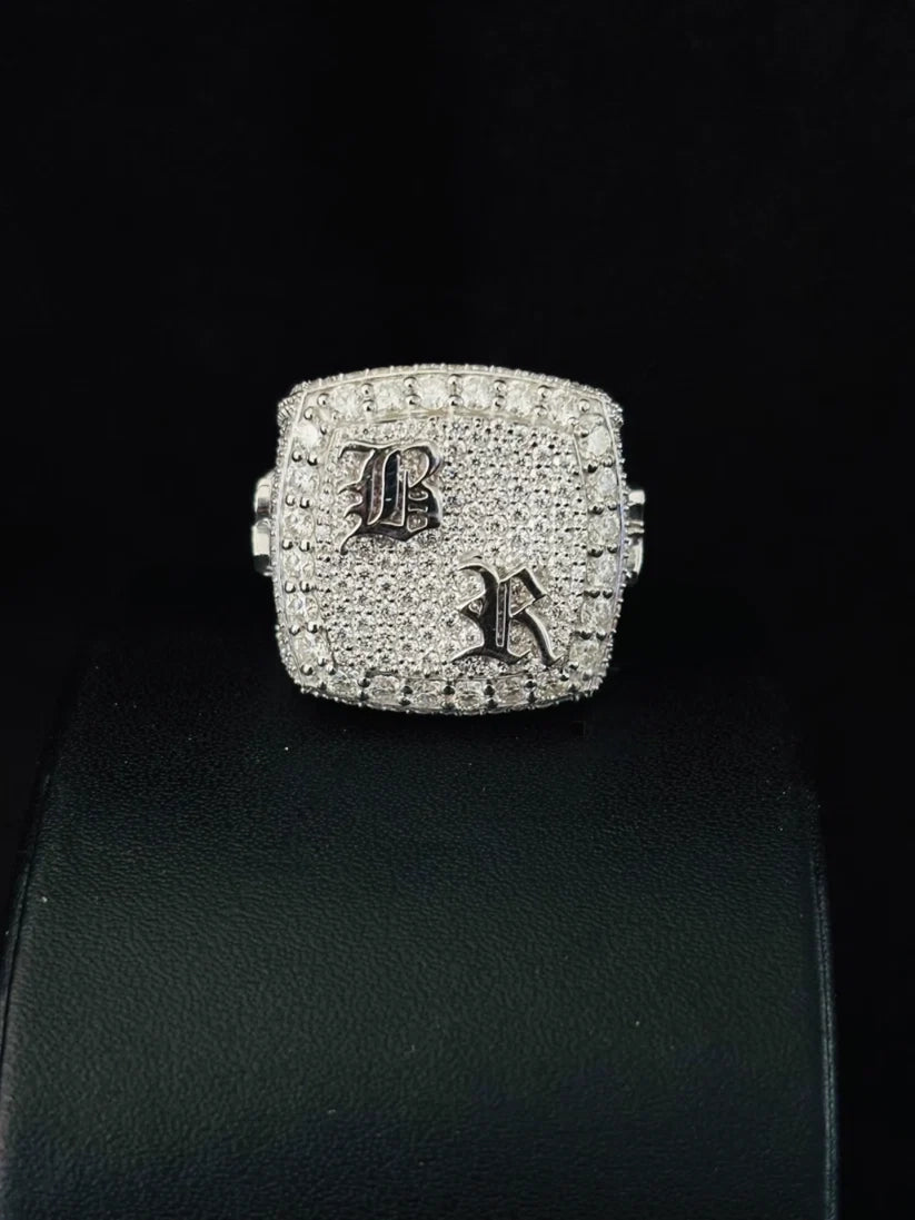 A square-shaped silver ring heavily paved with diamond, a gothic letter 'B' on one side and a gothic letter 'K' on the other