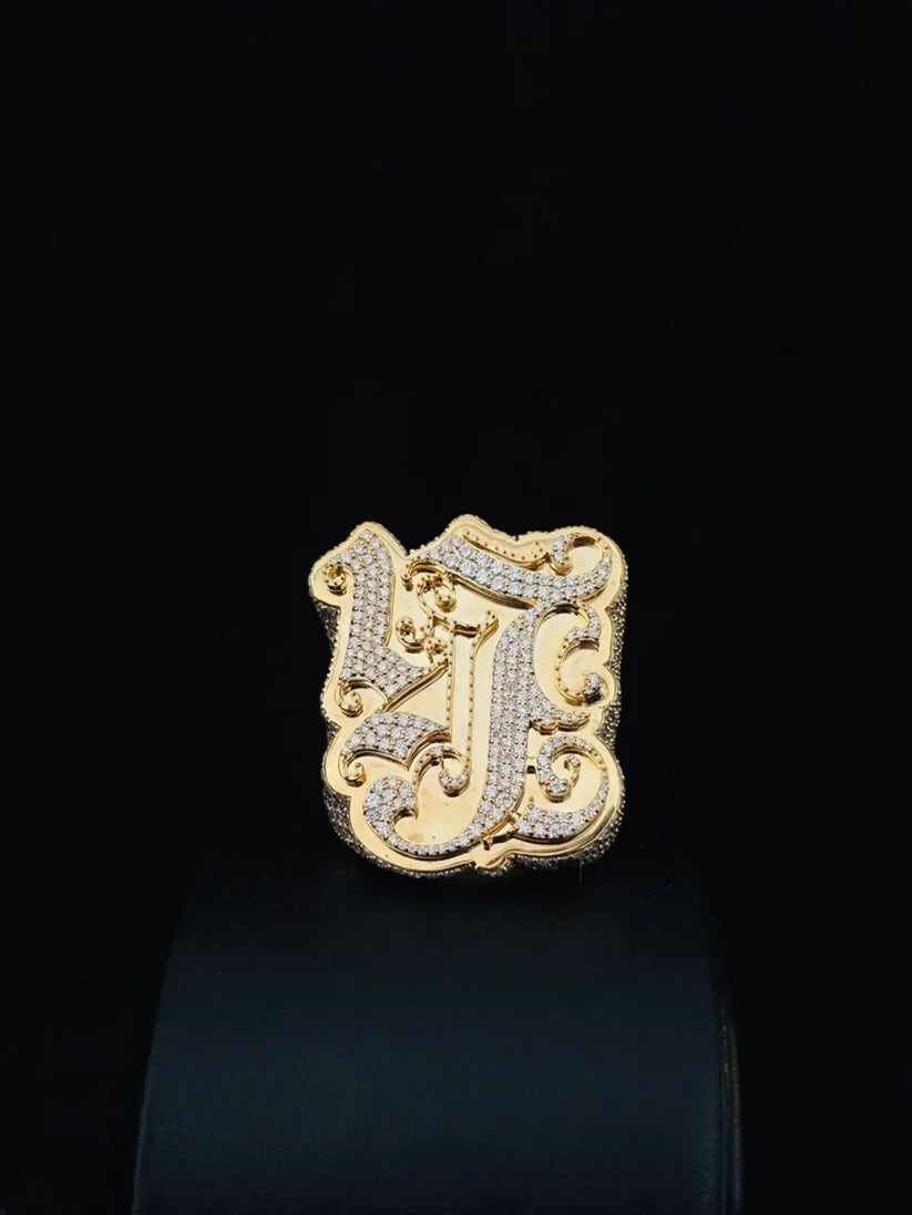 A gold plated initial 'J' ring paved with diamond, displayed on a black stand against a black background