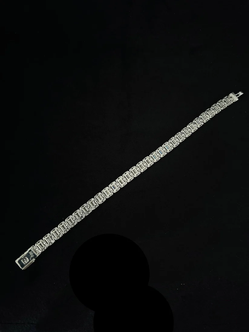 A silver tennis bracelet with rectangular clear stones, displayed diagonally on a black background