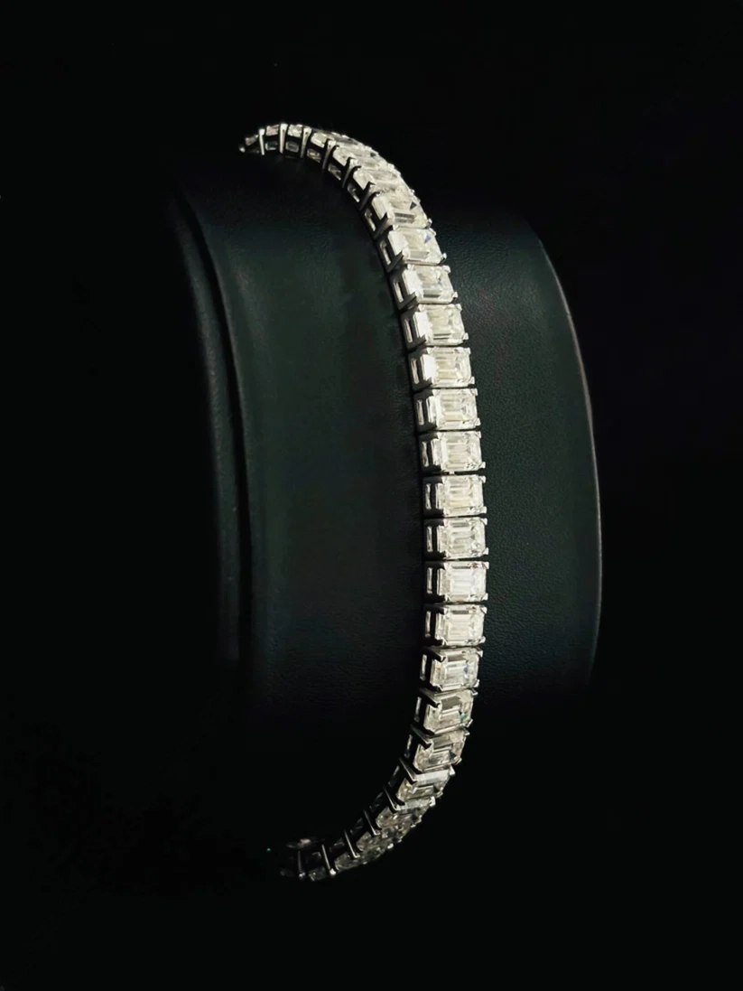 A side view silver tennis bracelet with emerald-cut diamond, displayed on a black cylindrical stand