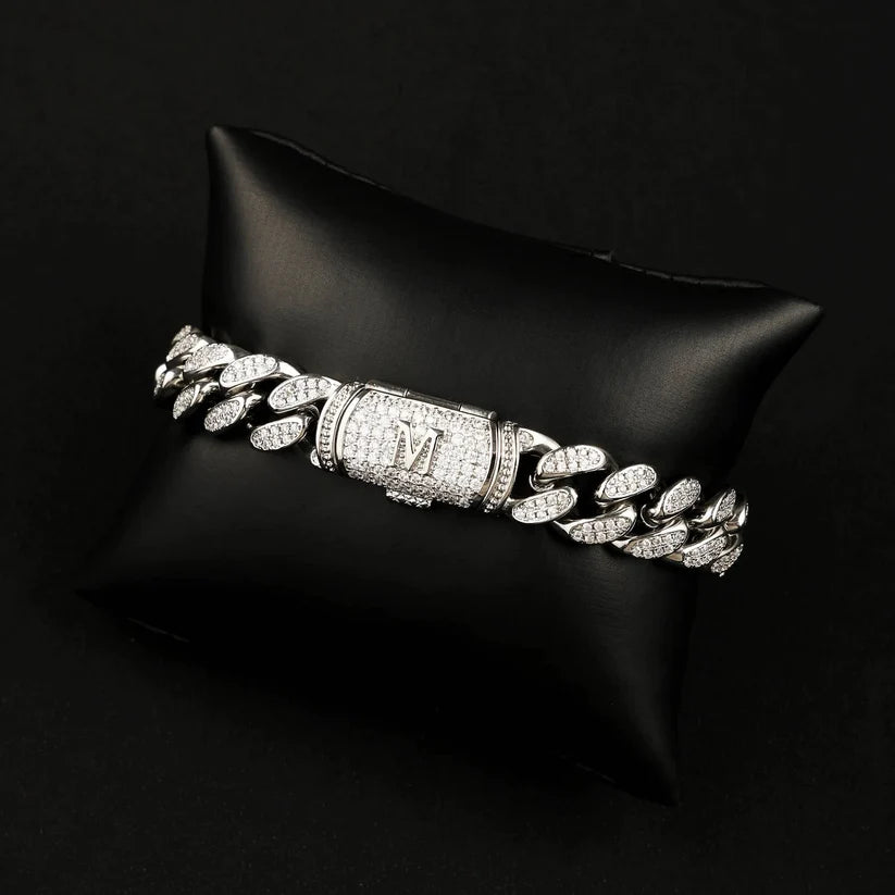 A top view silver Cuban link chain bracelet with a central clasp engraved with an 'M', displayed on a black satin pillow