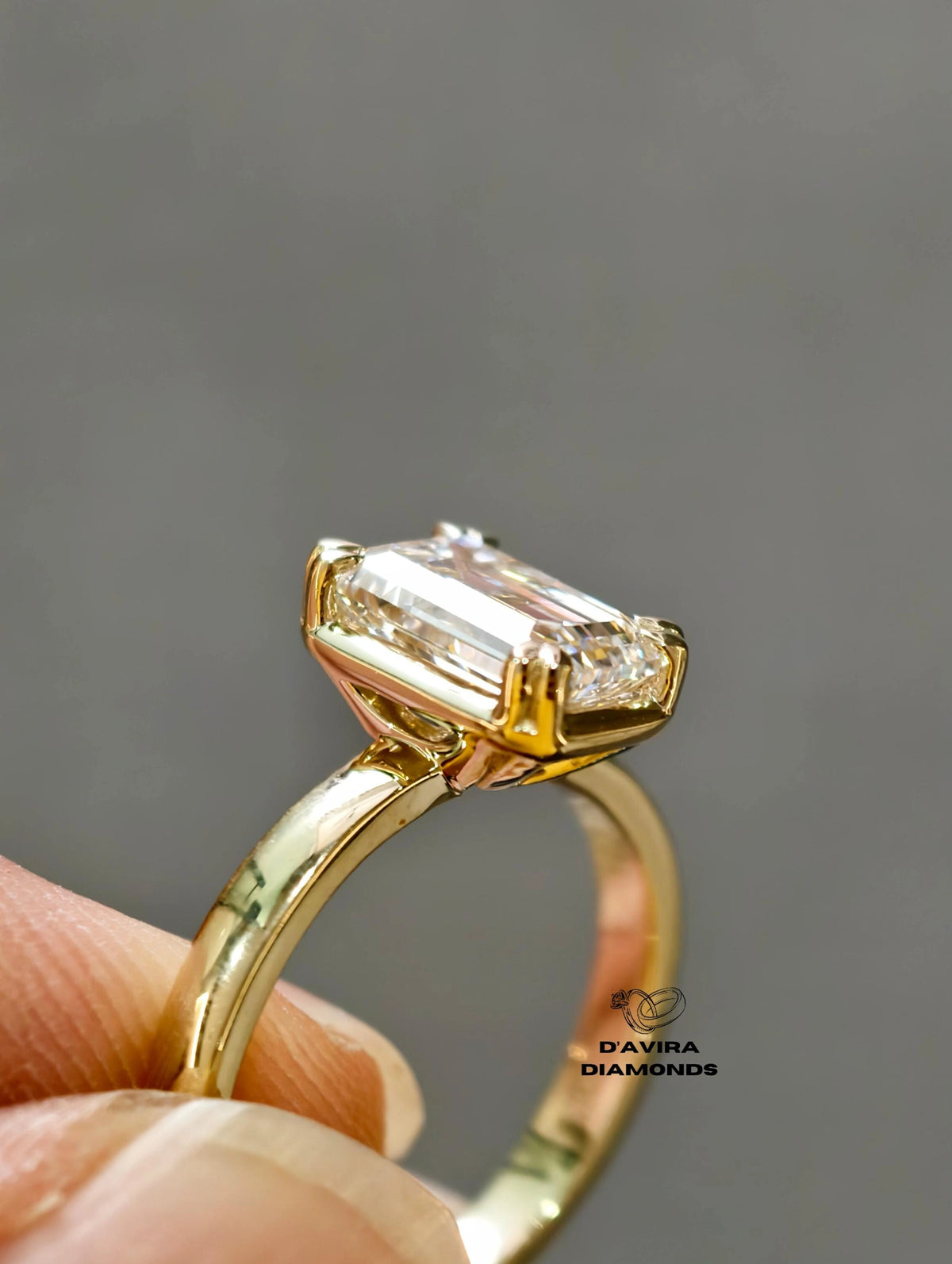A close-up of a gold ring with an emerald-cut diamond, held by finger