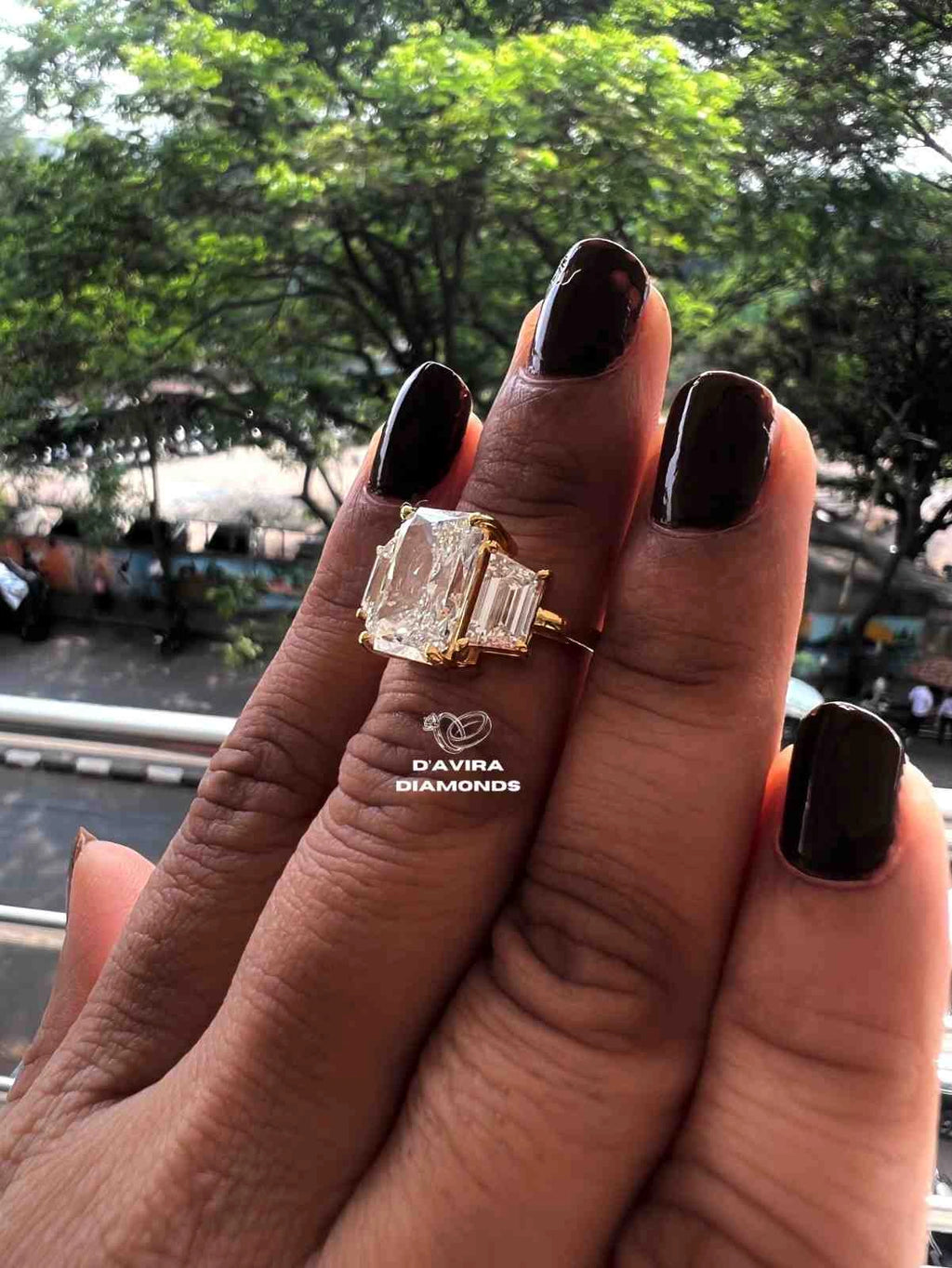 gold ring with a large rectangular clear gemstone and baguette side stones. The background shows a blurred outdoor street scene with green trees.