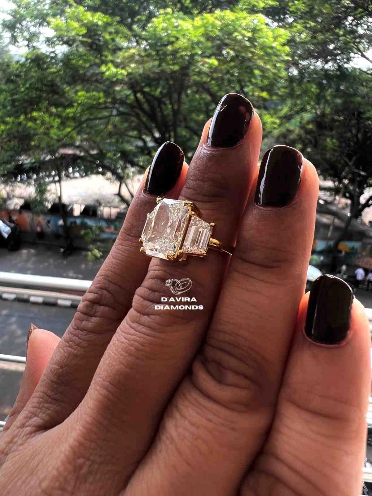 gold ring with a large rectangular clear gemstone and baguette side stones. The background shows a blurred outdoor street scene with green trees.