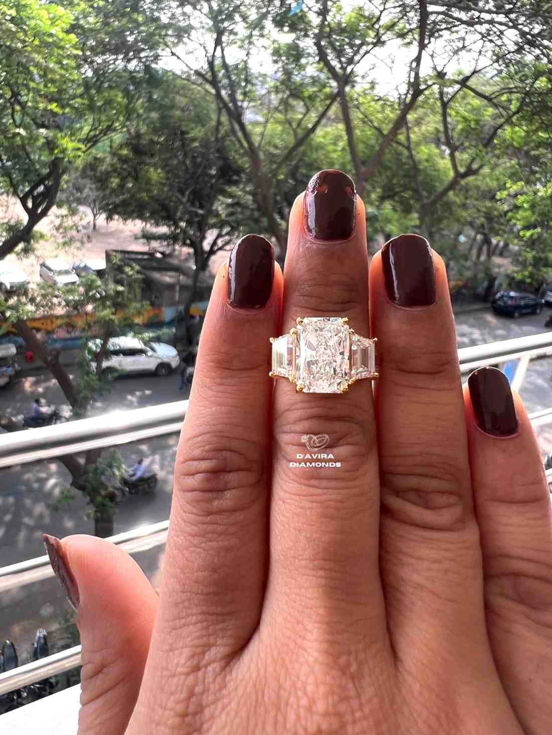 A close-up of a hand wearing a diamond ring with a central emerald-cut diamond and baguette side stones, set in a yellow gold band. The background shows an outdoor street scene with trees and parked cars.