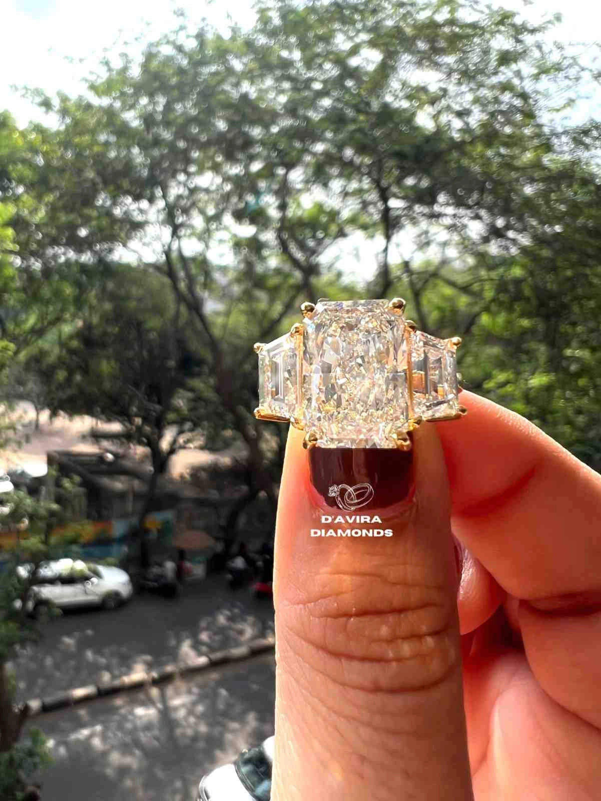 A hand holding a yellow gold ring with a large rectangular center stone and two smaller side stones, photographed outdoors with blurred trees and street background. The ring has a visible 'DAVIRA DIAMONDS' logo on the ring finger nail polish.