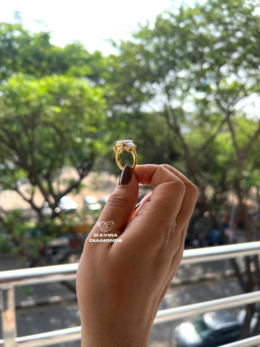 A hand holding a yellow gold ring with a large clear center stone and smaller side stones, photographed outdoors with a blurred background of green trees and a railing. The brand name 'DAVRA DIAMONDS' is watermarked near the hand.