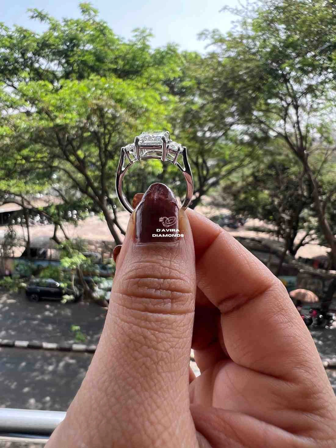 A silver ring with a large clear center stone held between fingers, with a blurred outdoor background of green trees and a street.